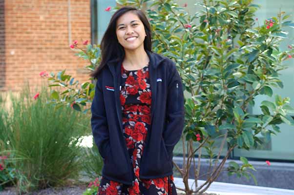 female student in red dress smiling outside in front of trees and building