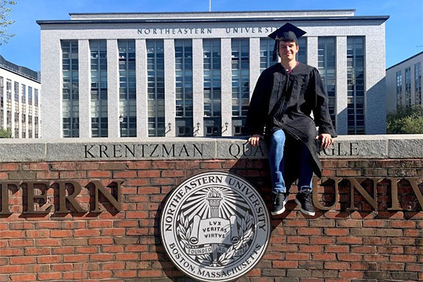 student sitting on Northeastern University wall in graduation attire